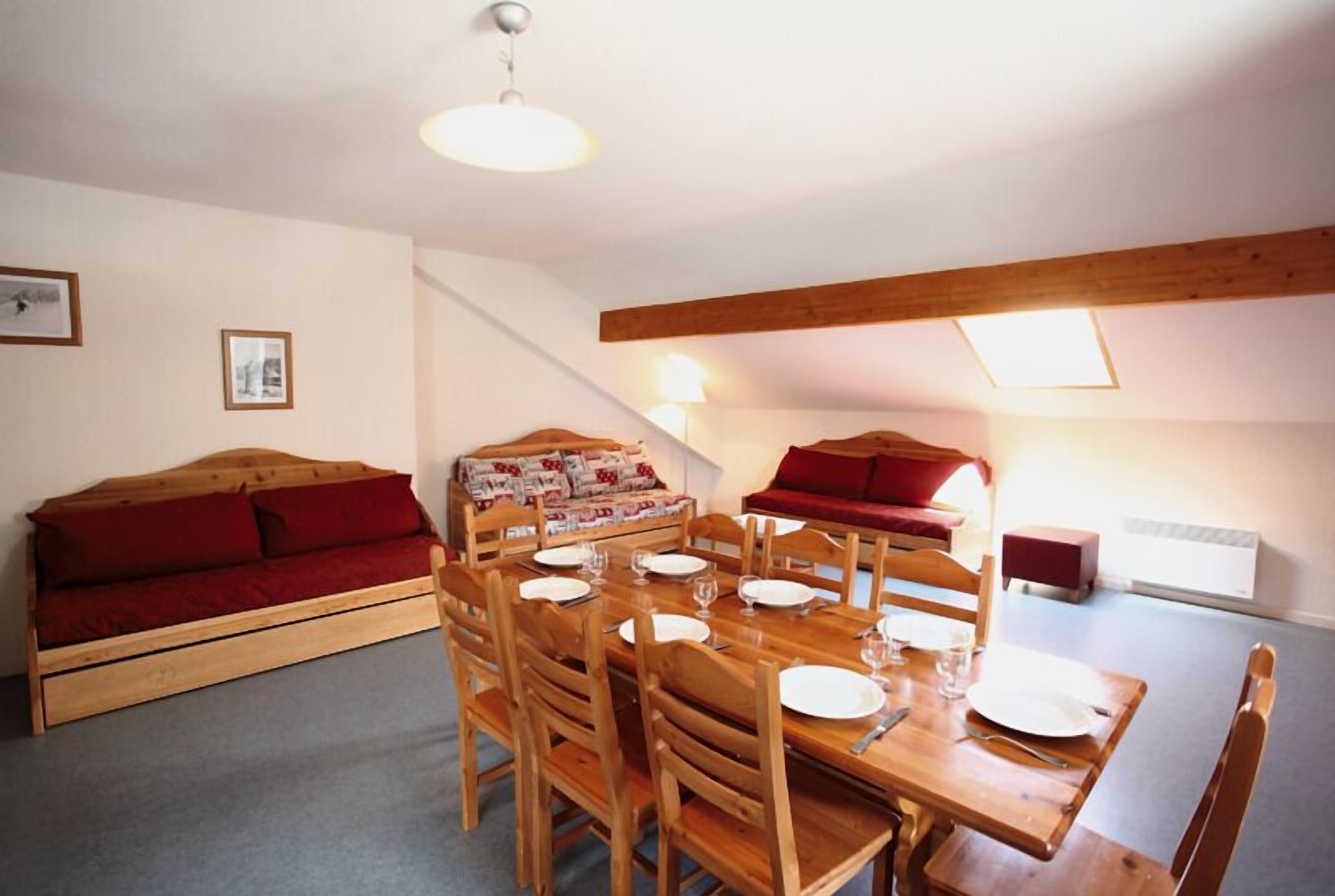 A dining area with a wooden table set for four, surrounded by wooden chairs. There is a red sofa with a patterned cover to the left and a red chair to the right. The room has a sloped ceiling with wooden beams and a skylight.