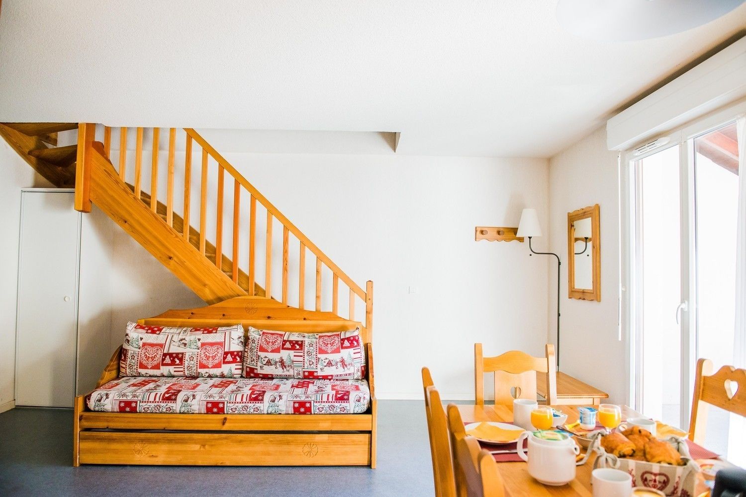 Une pièce chaleureuse avec un escalier en bois, un lit recouvert d’une couverture à motifs, une table à manger avec des aliments pour le petit-déjeuner et une fenêtre laissant entrer la lumière naturelle.