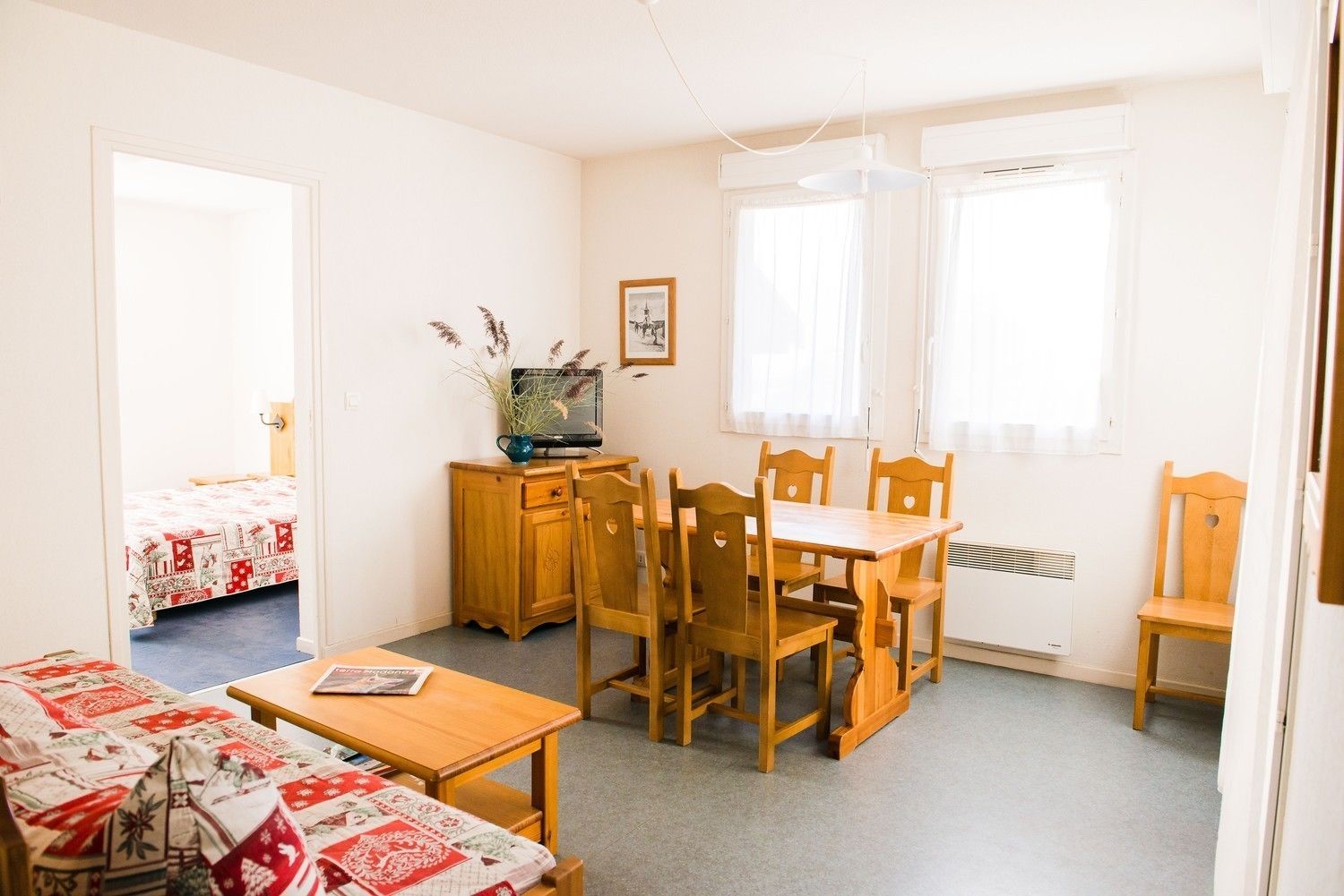 Una camera spaziosa con un tavolo da pranzo in legno, quattro sedie, un piccolo armadio, una televisione e un letto con copripiumino rosso e bianco.