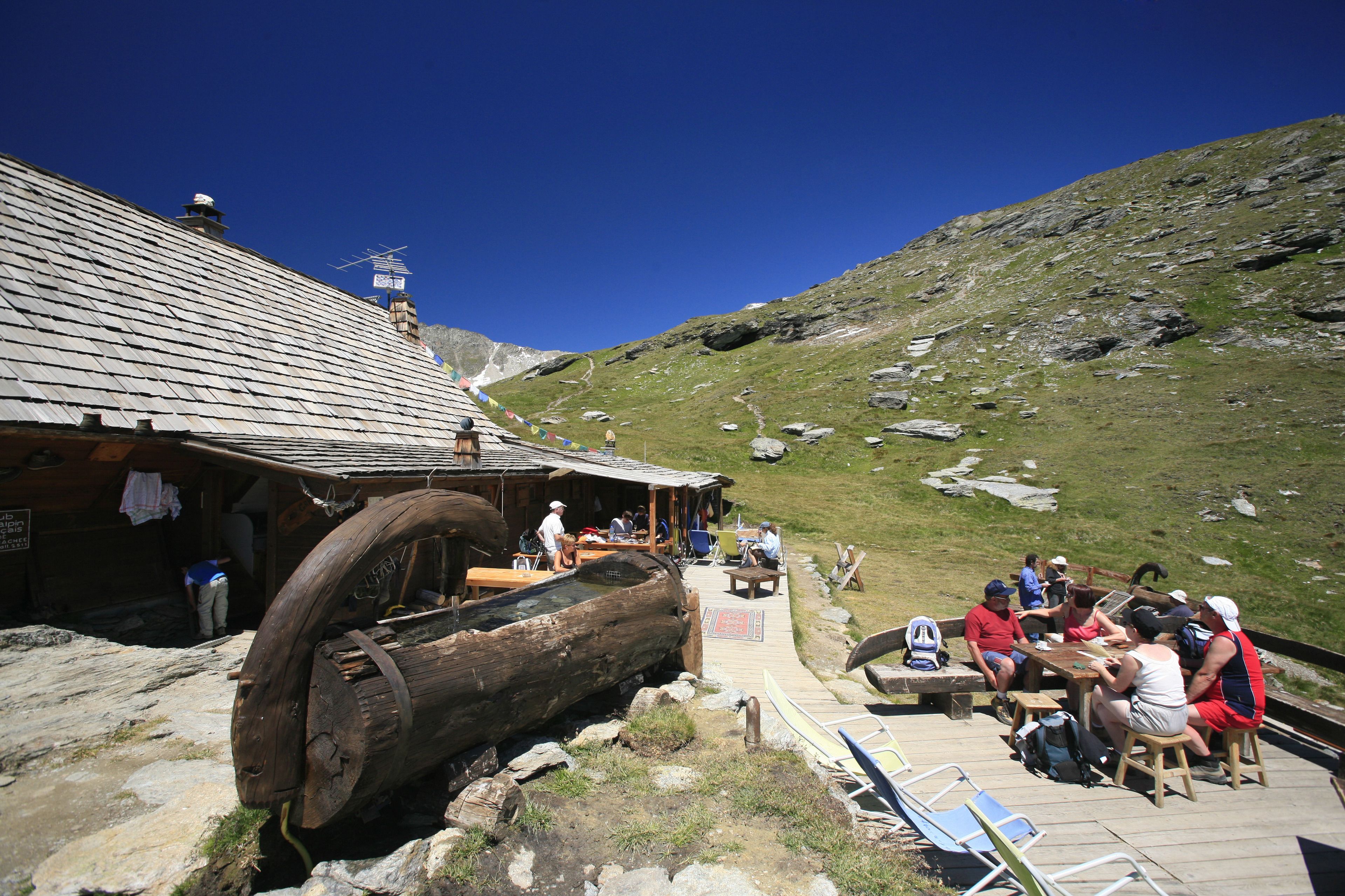 Un edificio rústico de madera con una gran rueda de leñe, una terraza de madera y un grupo de personas sentadas en una mesa.