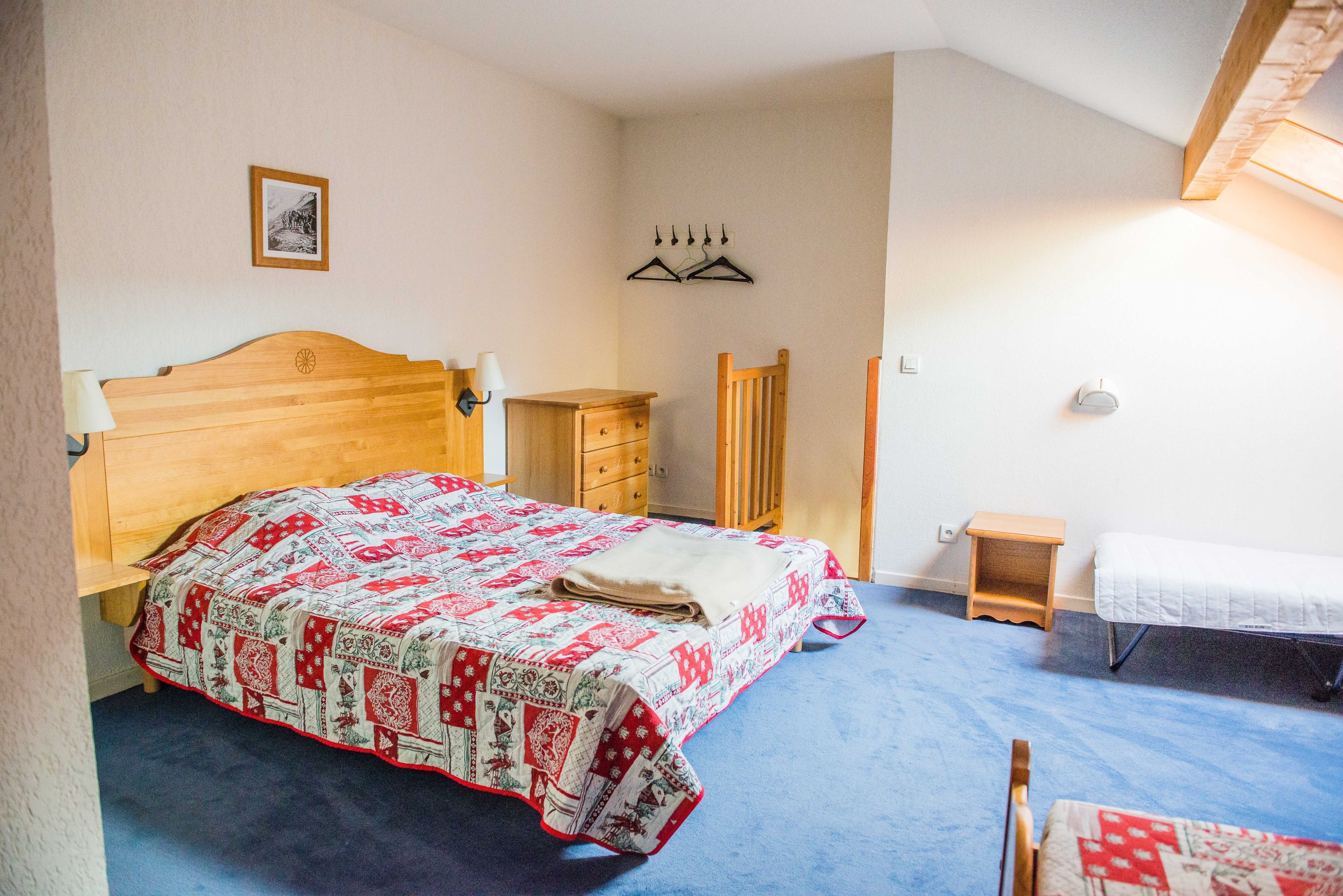A bedroom with a wooden bed, a small bedside table, a chair, a wardrobe, and a hanging coat rack.