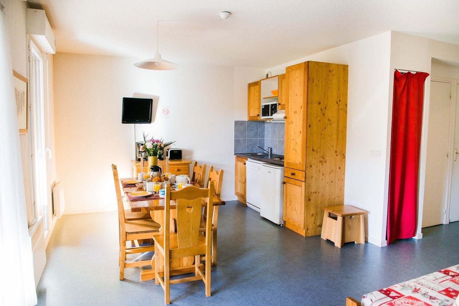 A compact kitchen with wooden cabinets, a dining table with chairs, a small refrigerator, and a red curtain.