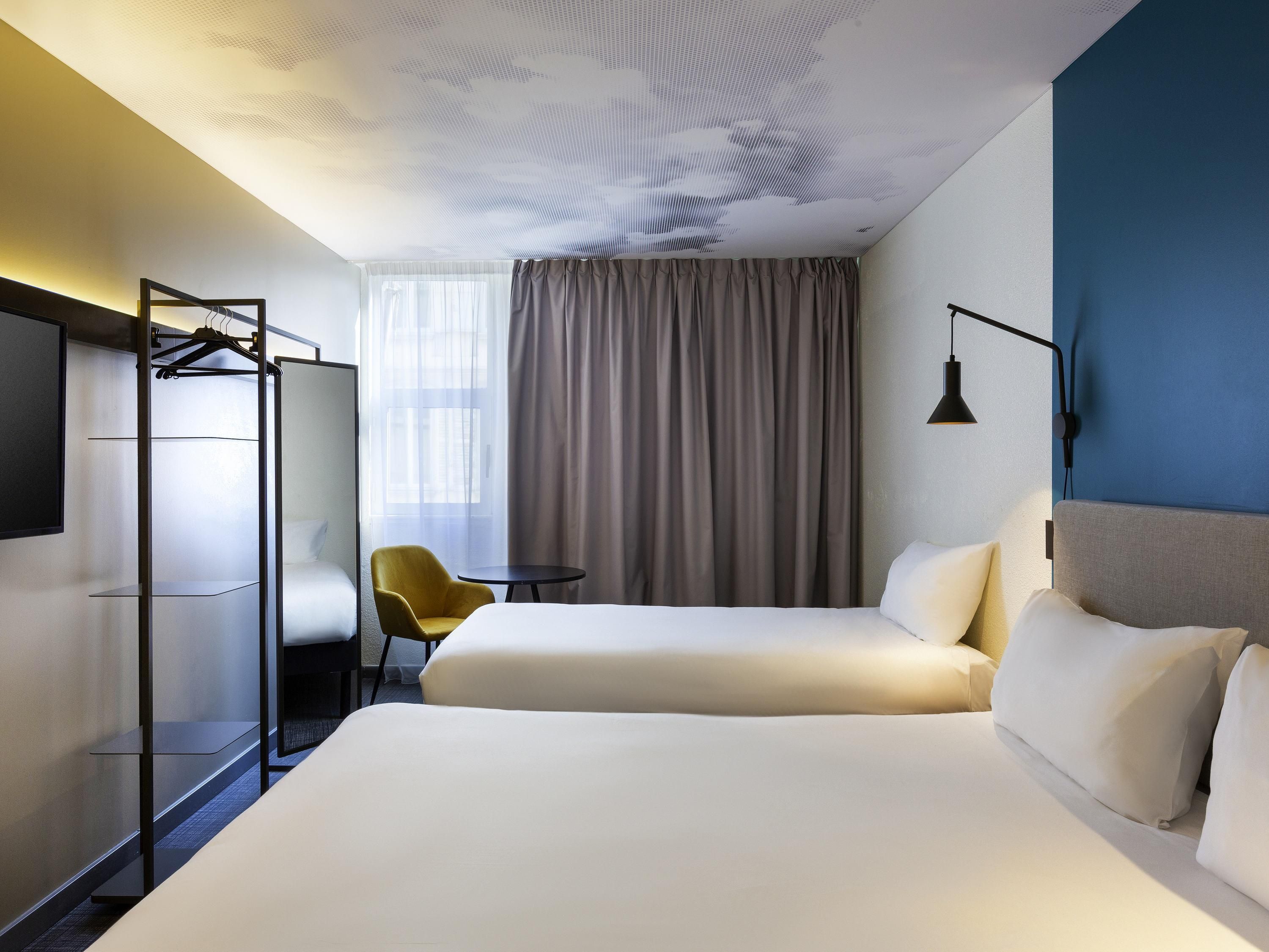 Habitació d'hotel amb un llit gran, una taula petita, una cadira groga, un armari i un televisor fixat a la paret.