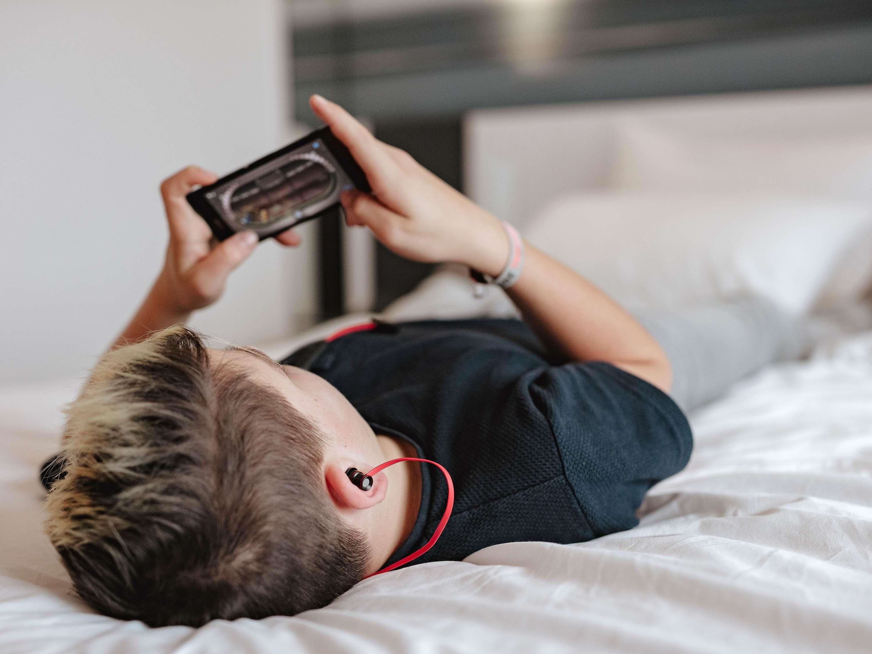 Uma pessoa deitada na cama com um smartphone, vestindo uma camisa preta e uma pulseira vermelha.