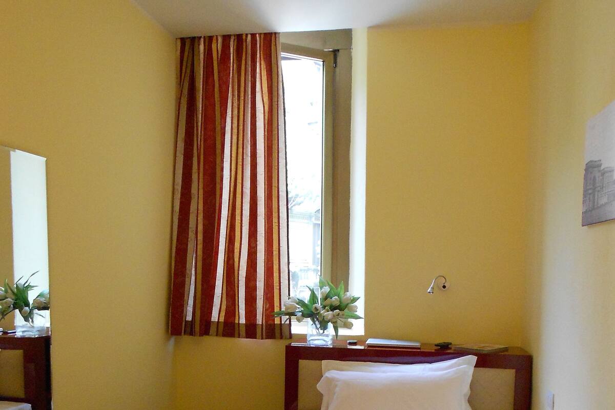 A hotel room with a bed, a chair, a window with curtains, a bedside table with a vase of flowers, and a wall-mounted painting.