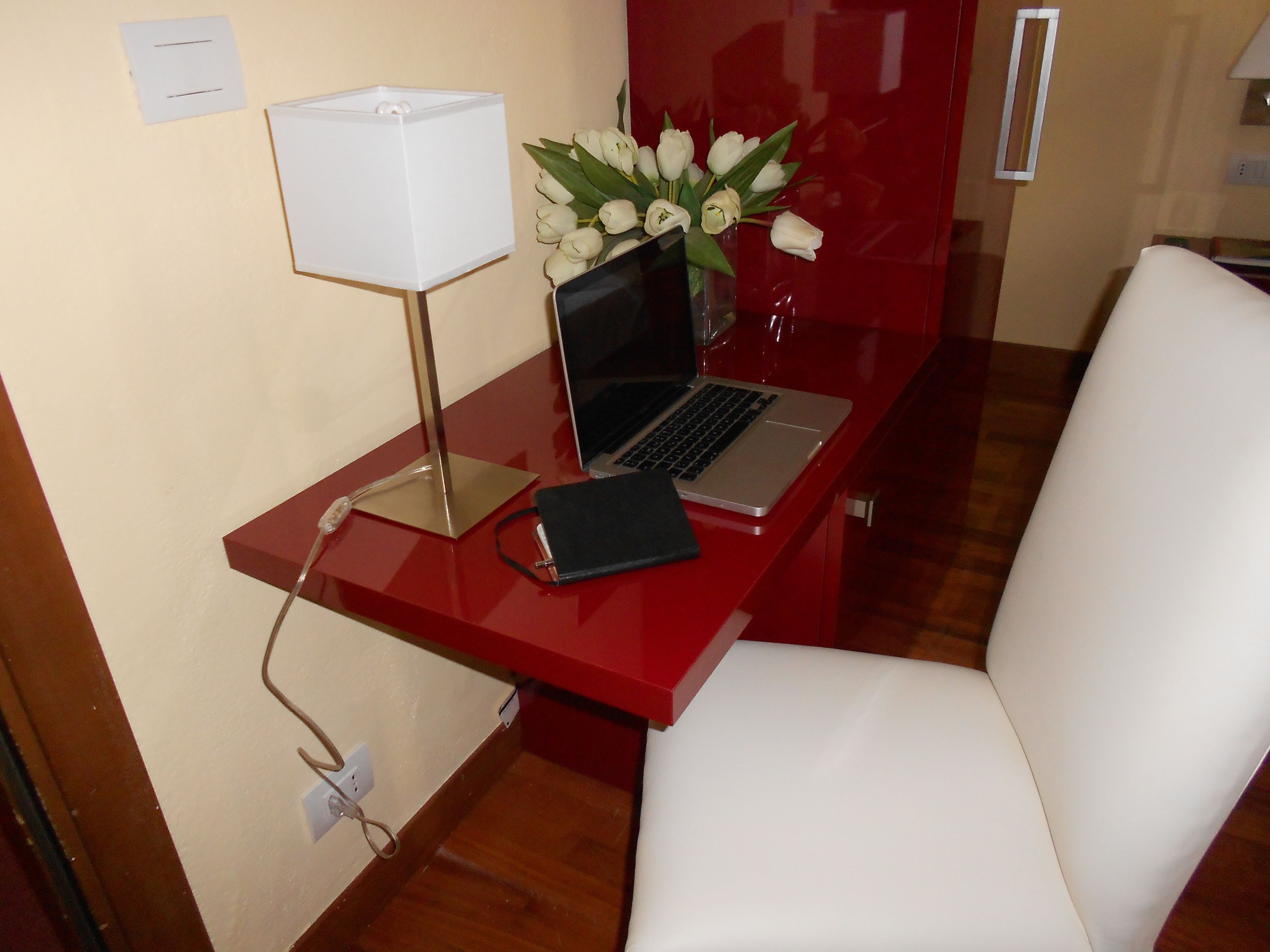 Un escritorio rojo con una computadora portátil, una libreta negra, una lámpara blanca y un florero con flores blancas.