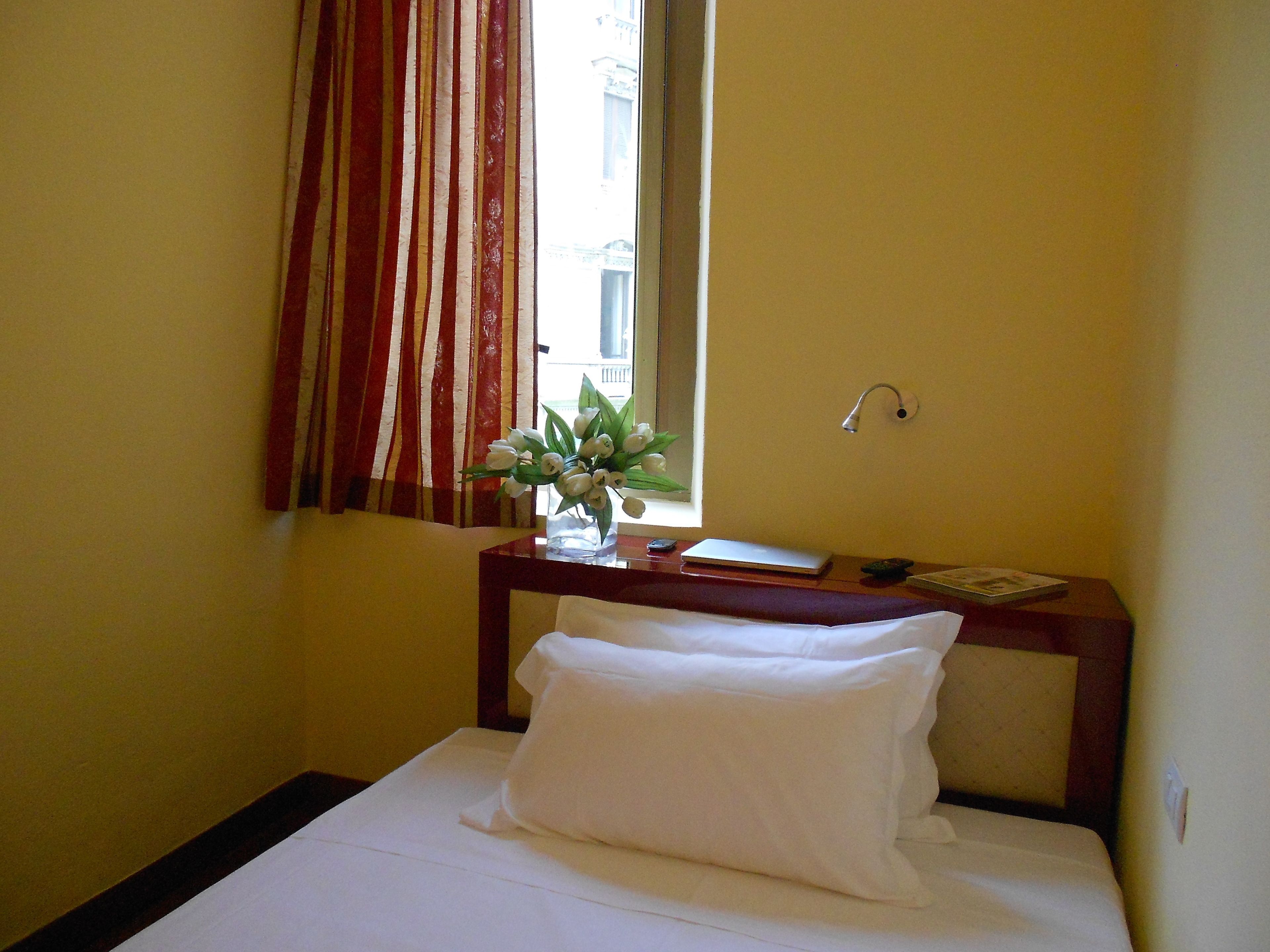 A hotel room with a bed, a bedside table with a vase of flowers, a window with curtains, and a nightstand with a lamp.