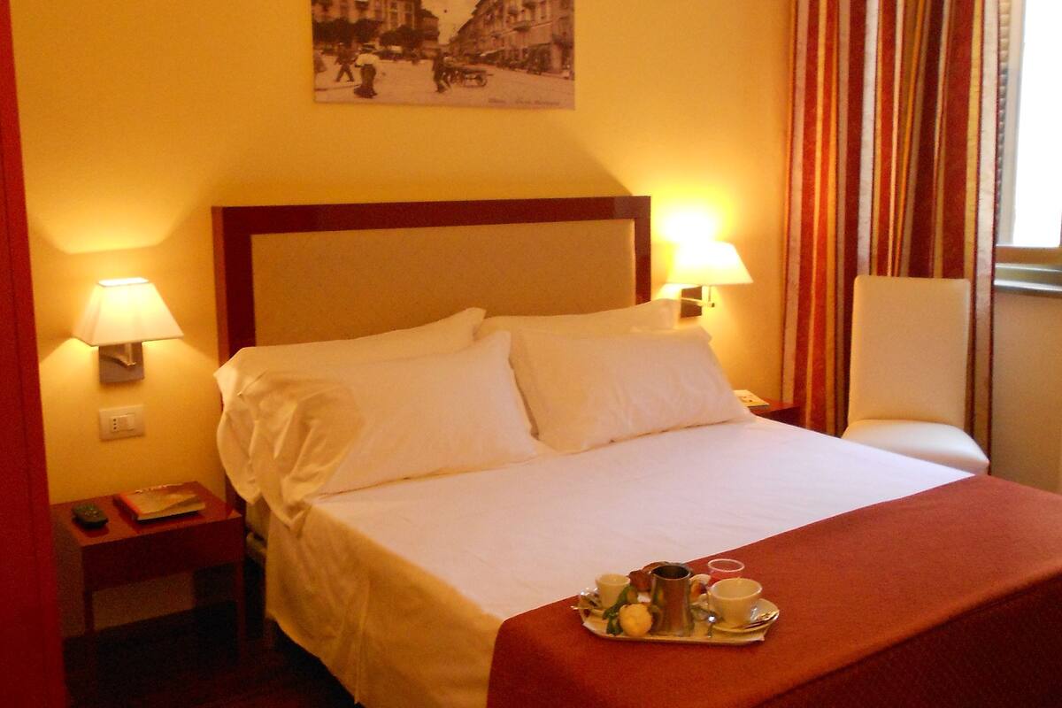 A hotel room with a bed, bedside lamps, a tray with breakfast items, and a wall-mounted picture.