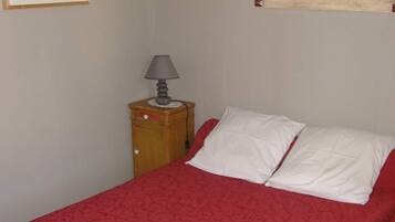 A single bed with a red bedspread, a wooden nightstand with a lamp, and a framed picture on the wall.
