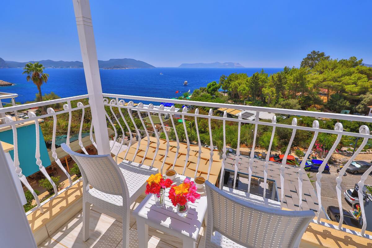 A balcony with white wicker chairs, a table with a vase of flowers, and a view of the sea and mountains.