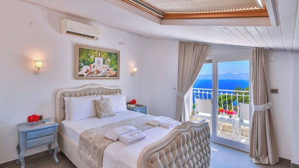 A bedroom with a large bed, a painting above the bed, a nightstand with a lamp, and a view of the sea through the balcony doors. - Linda Beach Class Hotel - Boutique Class (Kas)