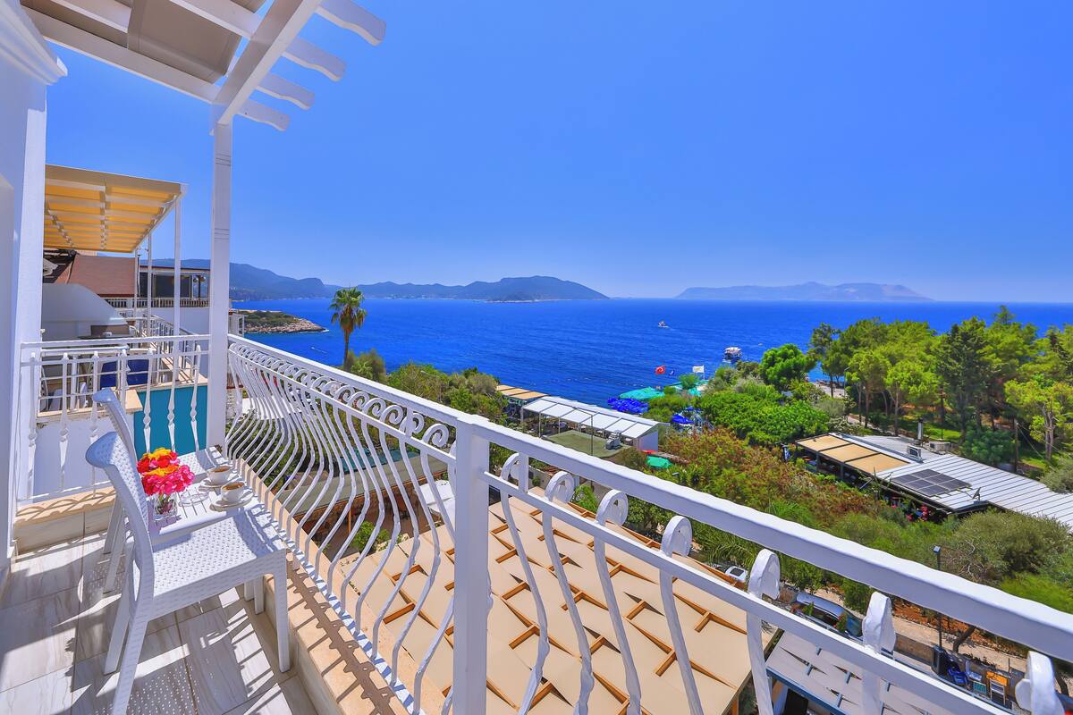 A balcony with a view of the sea, a palm tree, and a beach with umbrellas.
