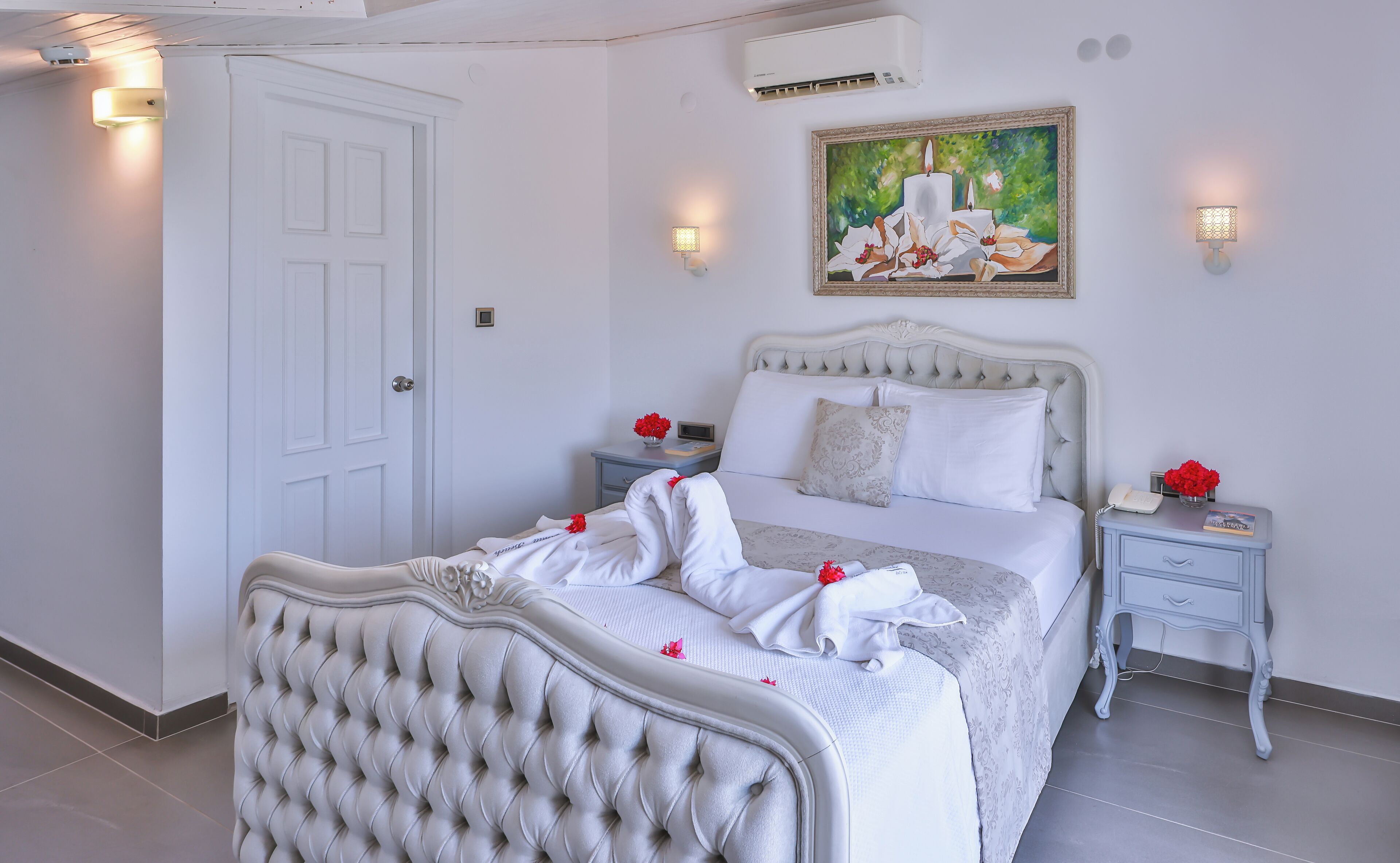 A neatly arranged bedroom with a large bed, two bedside tables, a wall-mounted air conditioner, and a painting above the bed.