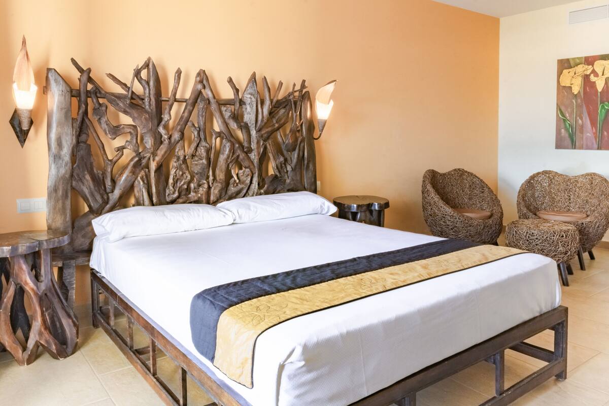 Ein Bett mit Holz-Kopfteil, ein runder Nachttisch, zwei Korbstühle und ein Bild an der Wand.