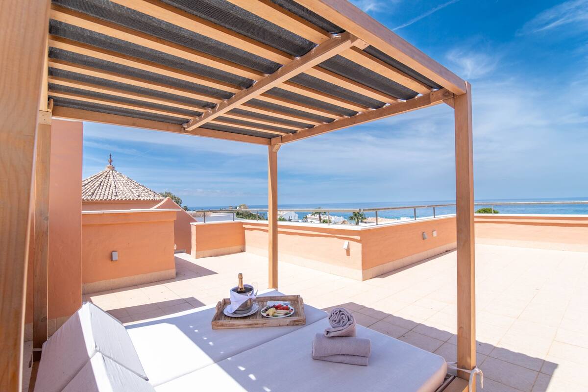 Eine Dachterrasse mit einer Holz-Pergola, einem Esstisch mit einer Flasche und Früchten sowie einem Handtuch.