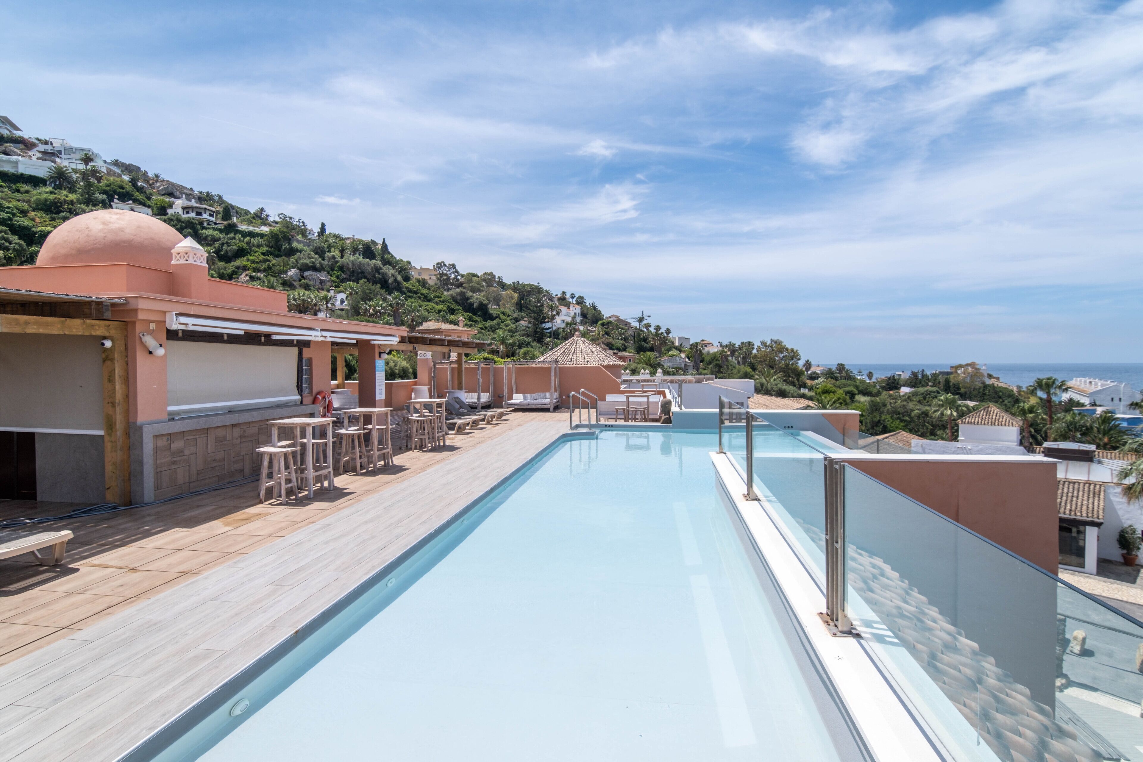 Piscina en la azotea con sillones de descanso y una terraza de madera, con vistas a una colina con edificios y un cielo despejado.