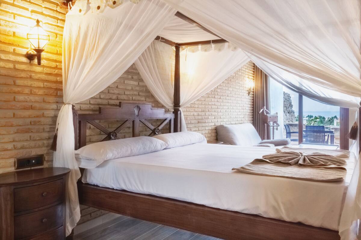 Ein geräumiges Schlafzimmer mit einem großen Bett, einem Holz-Kopfteil, einem Nachttisch und Blick ins Freie durch ein großes Fenster.
