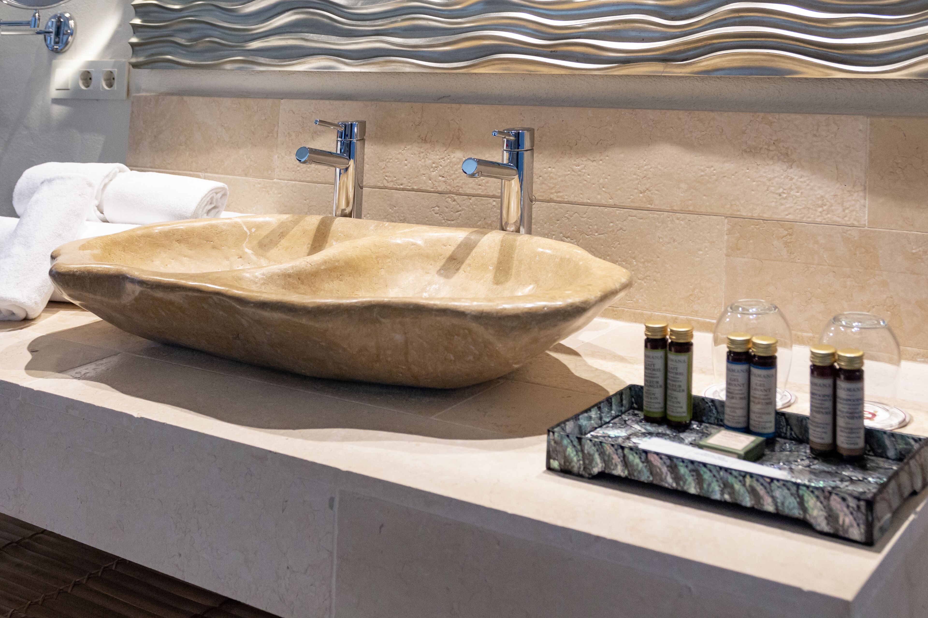 Une salle de bain dotée d’un grand lavabo de forme ovale, de deux robinets chromés et d’un comptoir en marbre. Plusieurs flacons de produits de toilette sont posés sur le comptoir.