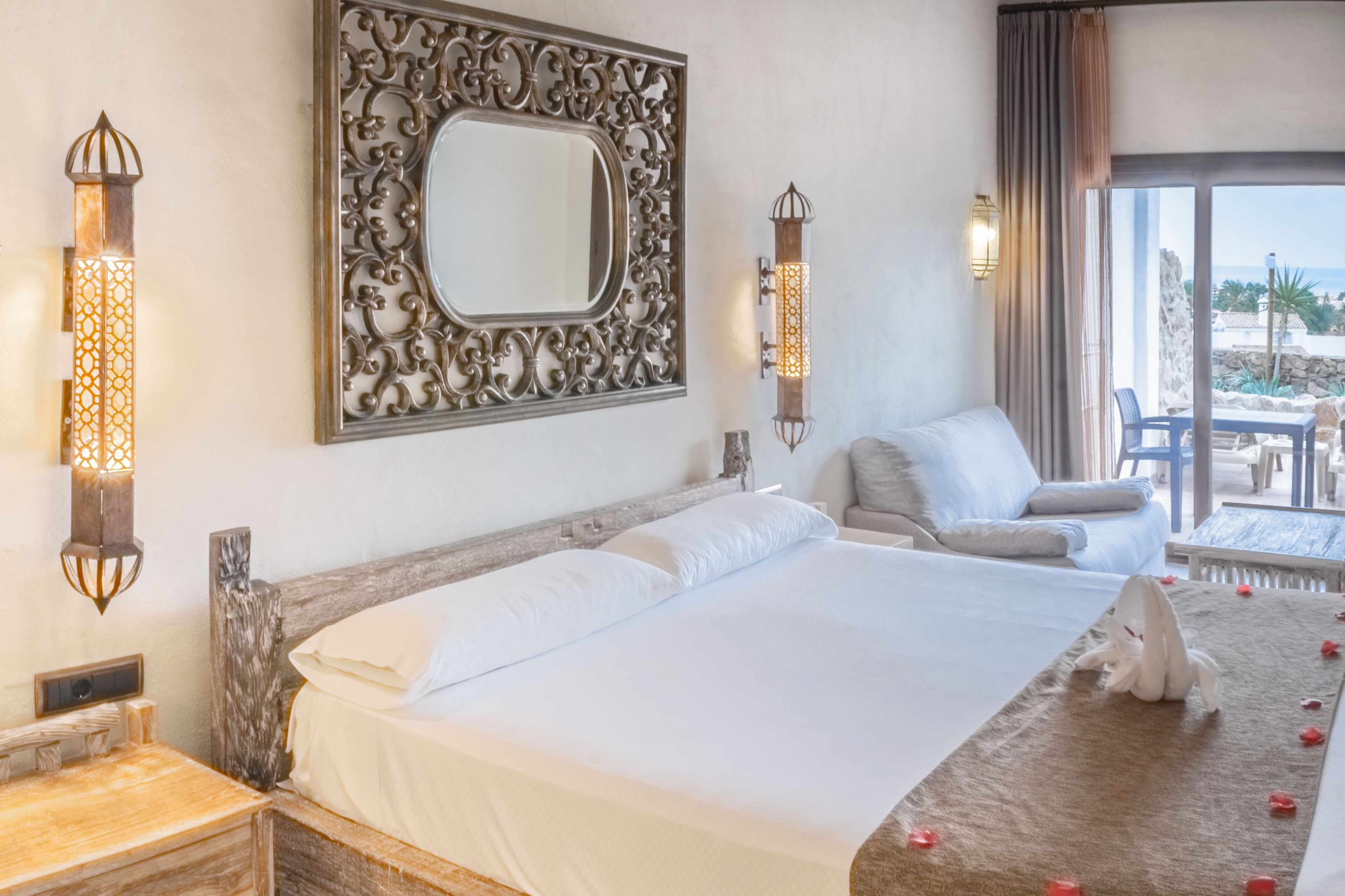 Quarto de hotel com uma cama grande, um espelho decorativo, arandelas na parede e vista para a praia pela janela.