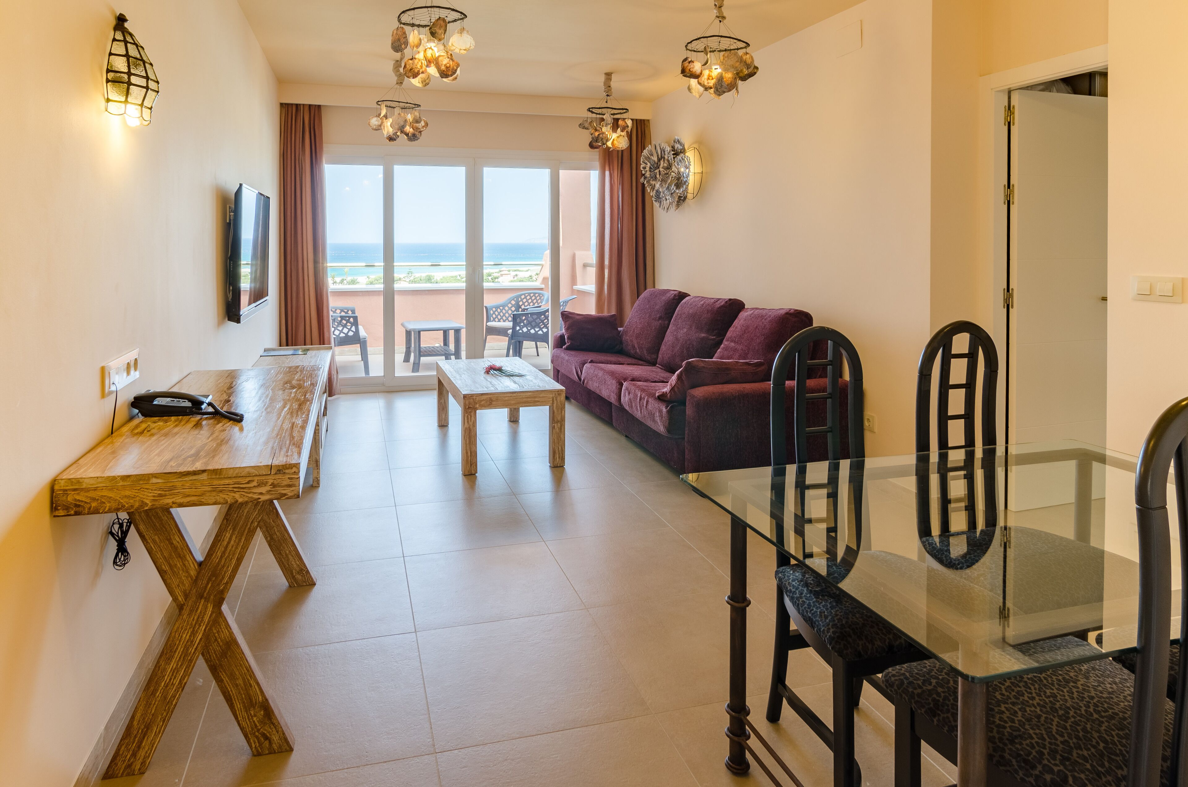 Una sala de estar con sofá, mesa de comedor de vidrio y mesa de centro de madera. Hay un televisor montado en la pared y una vista al océano a través de la puerta corrediza de cristal.