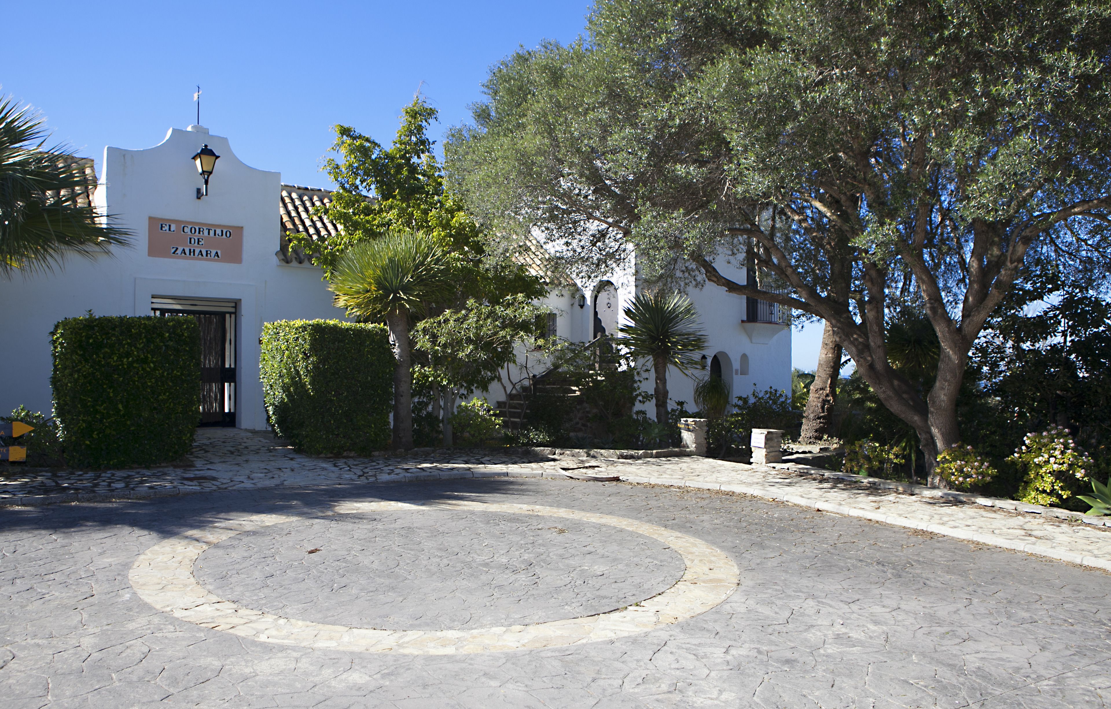 A courtyard with a circular stone pattern, a white building with a sign reading "El Cortijo de Zahara", and surrounding greenery.