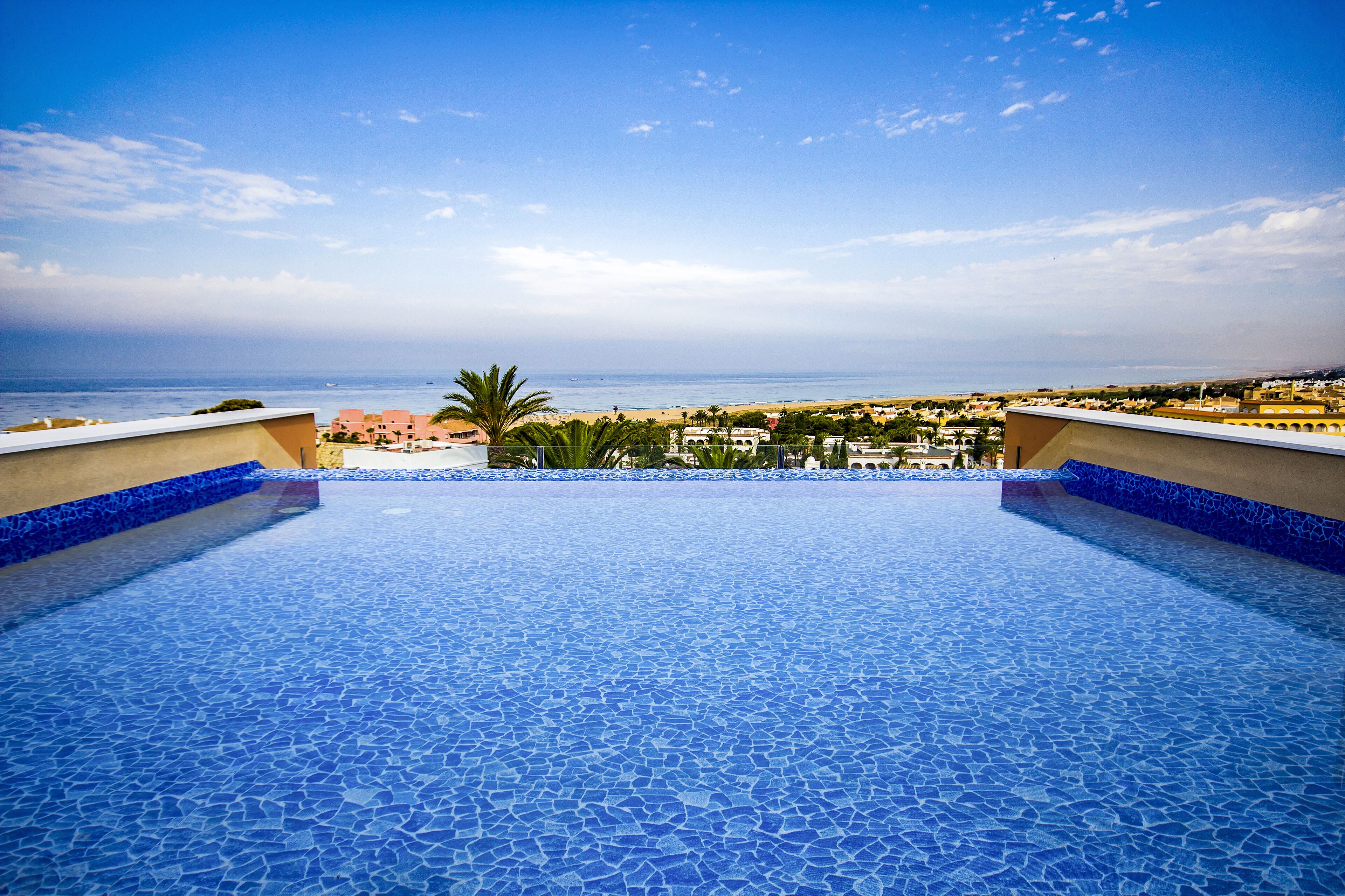 A rooftop pool with a blue mosaic floor, overlooking a beach and residential area.