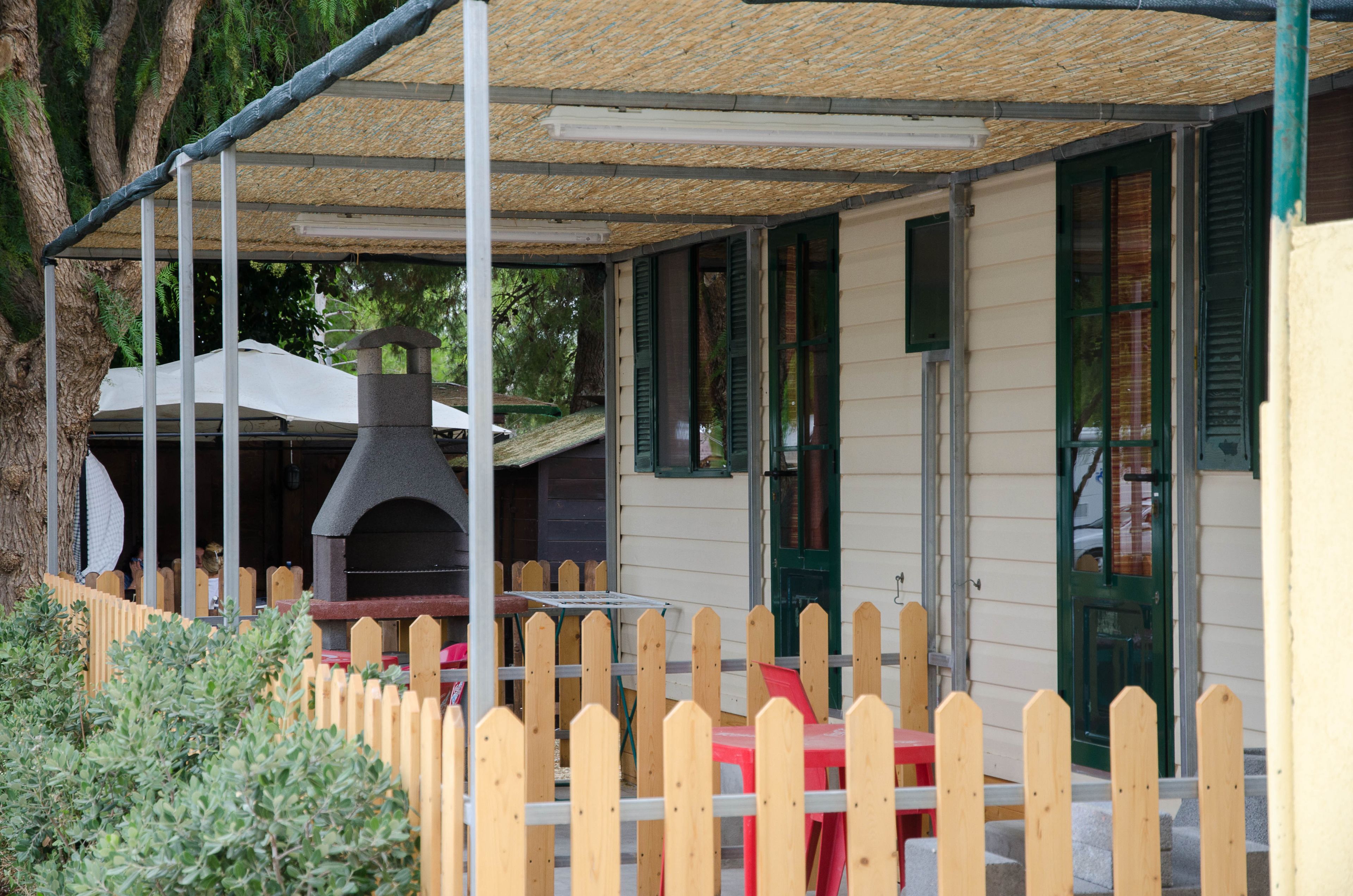 A covered outdoor seating area with a wooden fence, a barbecue grill, and a table with a red chair.
