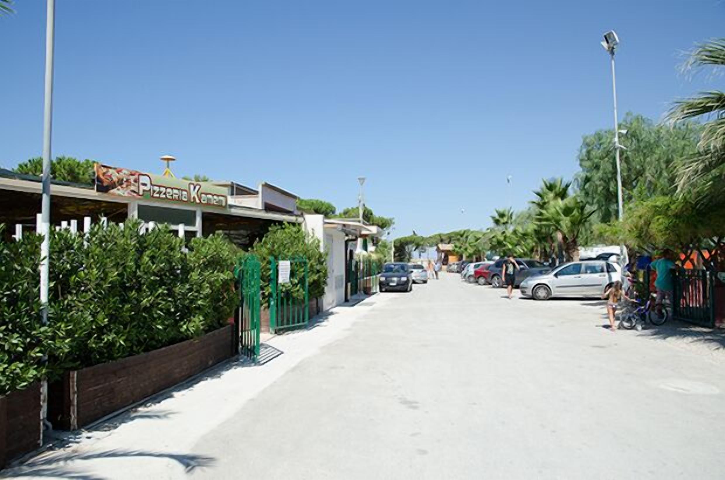 A street with a pizzeria named "Pizzeria Kamen" on the left, parked cars, and a clear blue sky.
