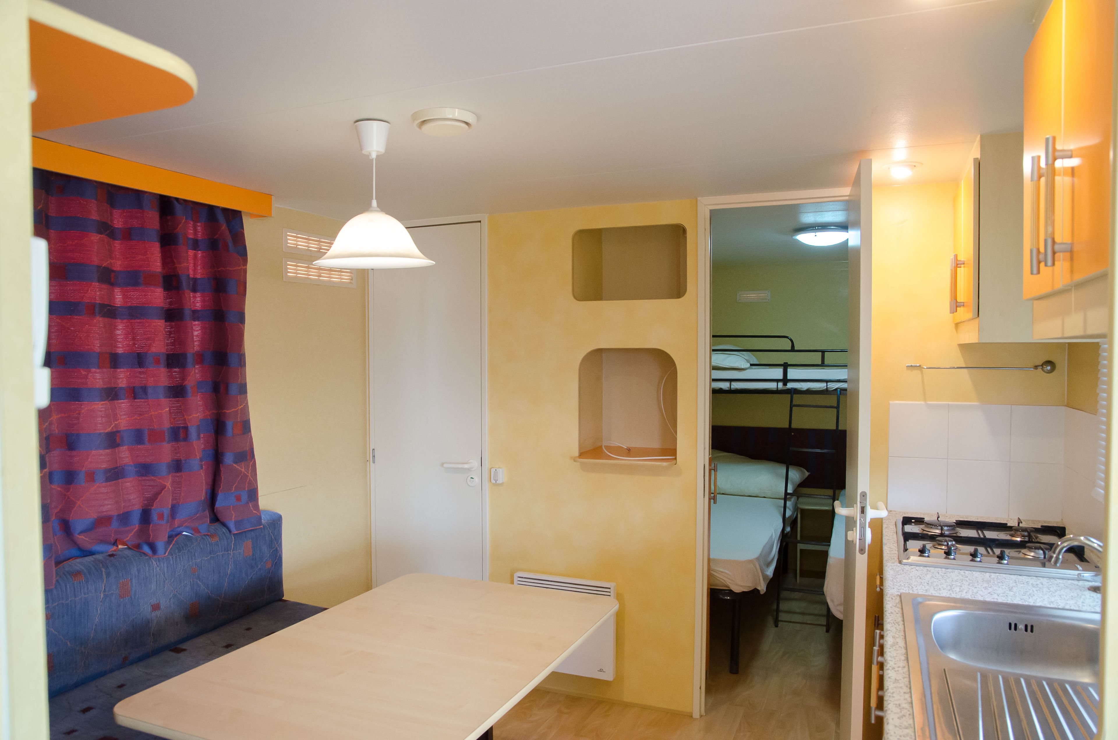 A compact room with a bunk bed, a small kitchenette, a table, and a window with curtains.
