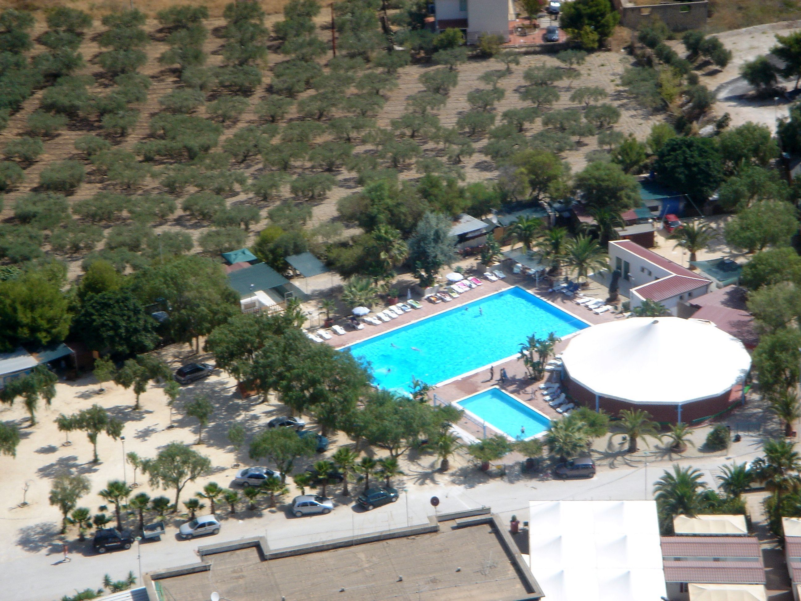 Luftaufnahme eines Resorts mit einem zentralen Schwimmbecken, umgeben von Palmen und Liegestühlen.