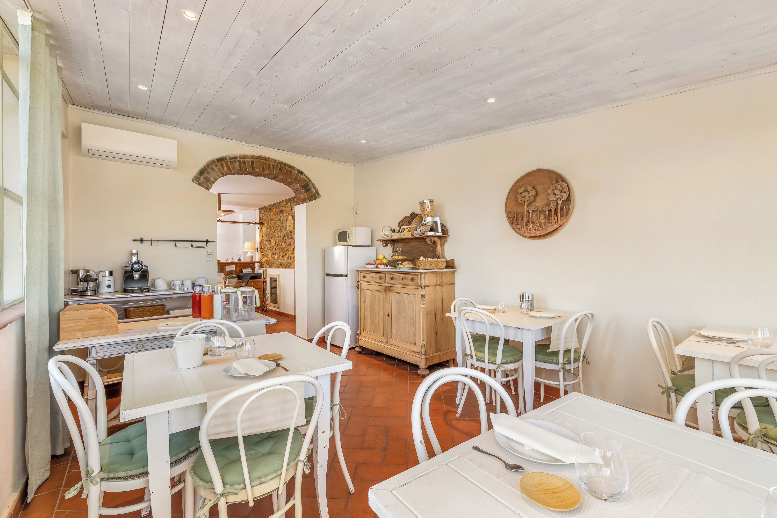 A dining area with white tables and chairs, a kitchen in the background, and a rustic wooden cabinet.