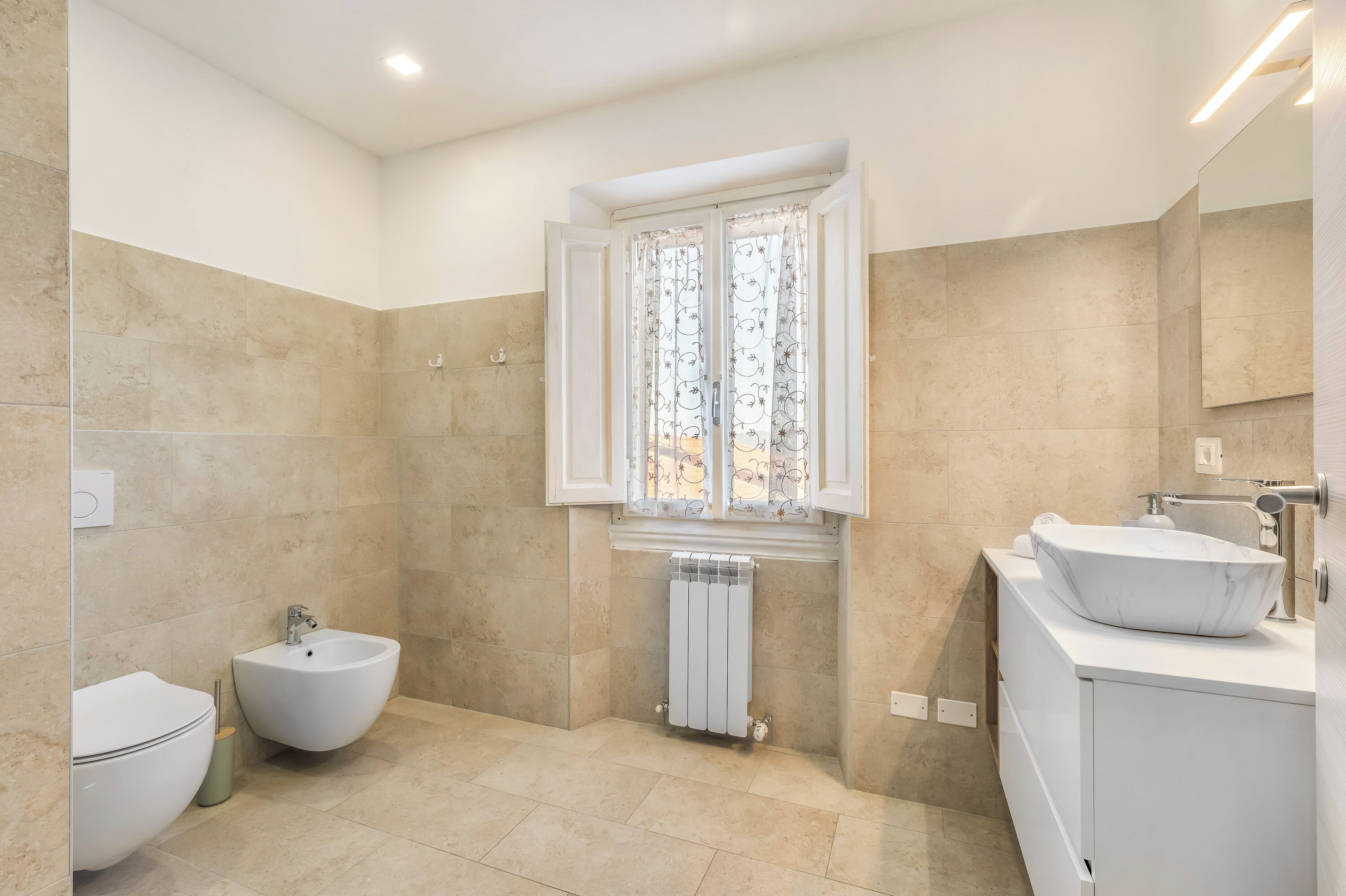 A bathroom with a bidet, a toilet, a sink with a marble basin, a window with patterned curtains, and a radiator.