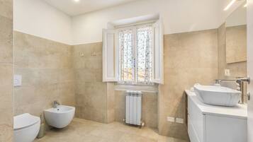 A bathroom with a bidet, a toilet, a sink with a marble basin, a window with patterned curtains, and a radiator.