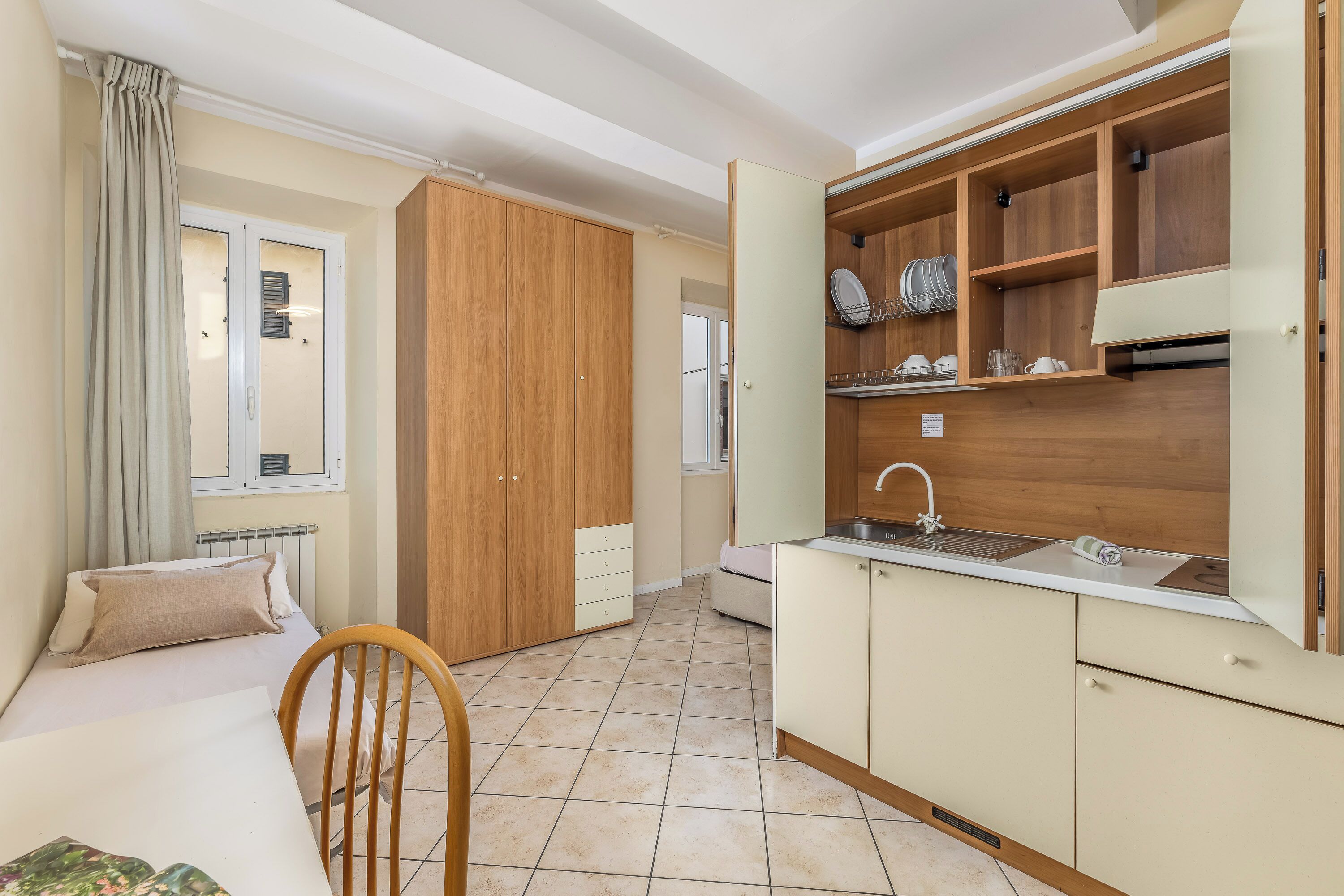 Quarto compacto com cama, guarda-roupa de madeira, cozinha compacta com pia e janela com cortinas.