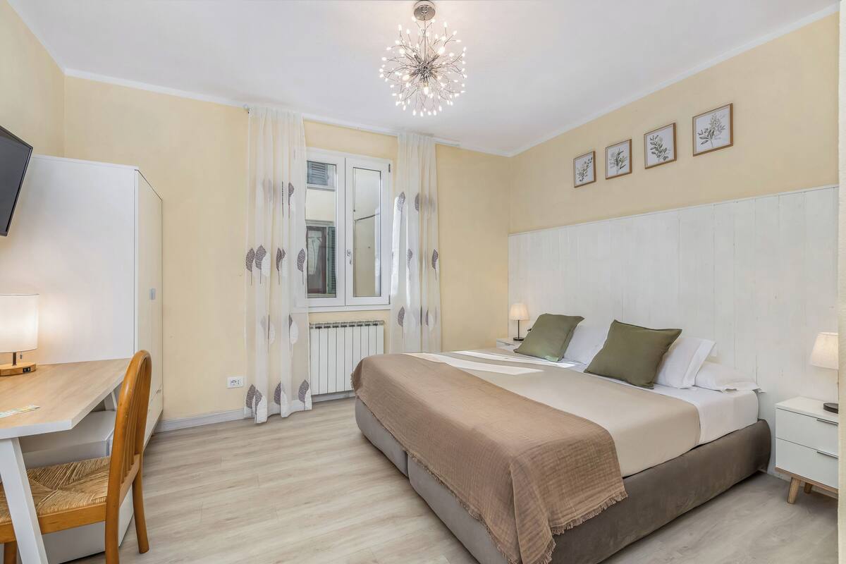 A bedroom with a bed, bedside tables, a desk, a chair, a window with curtains, and a ceiling light fixture.