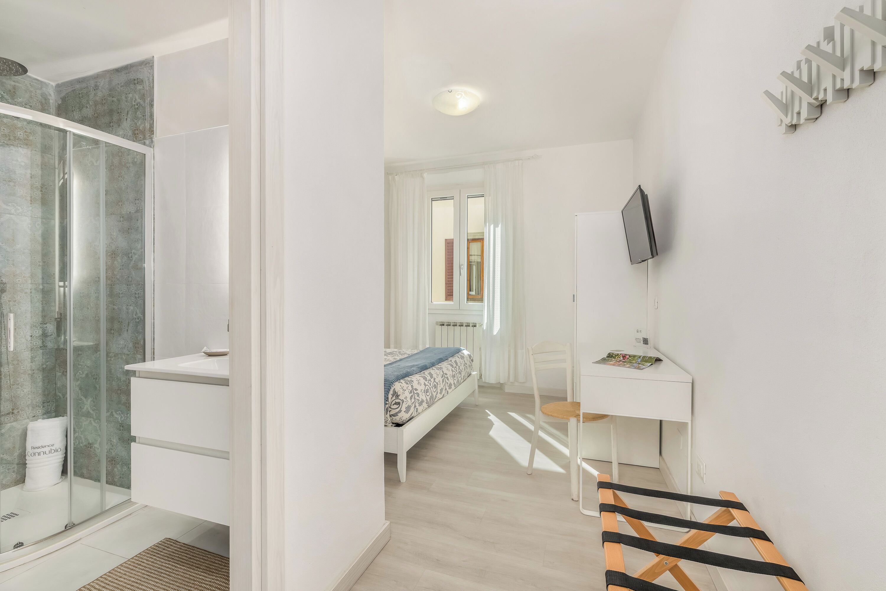 Un dormitorio moderno con cabina de ducha de vidrio, una cama con colcha estampada, un escritorio con silla y un televisor montado en la pared.