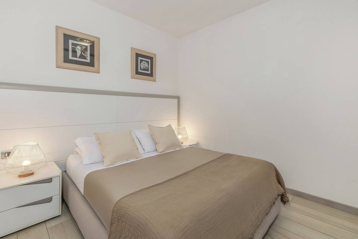 A neatly made bed with a beige blanket, a white headboard, and two bedside lamps.