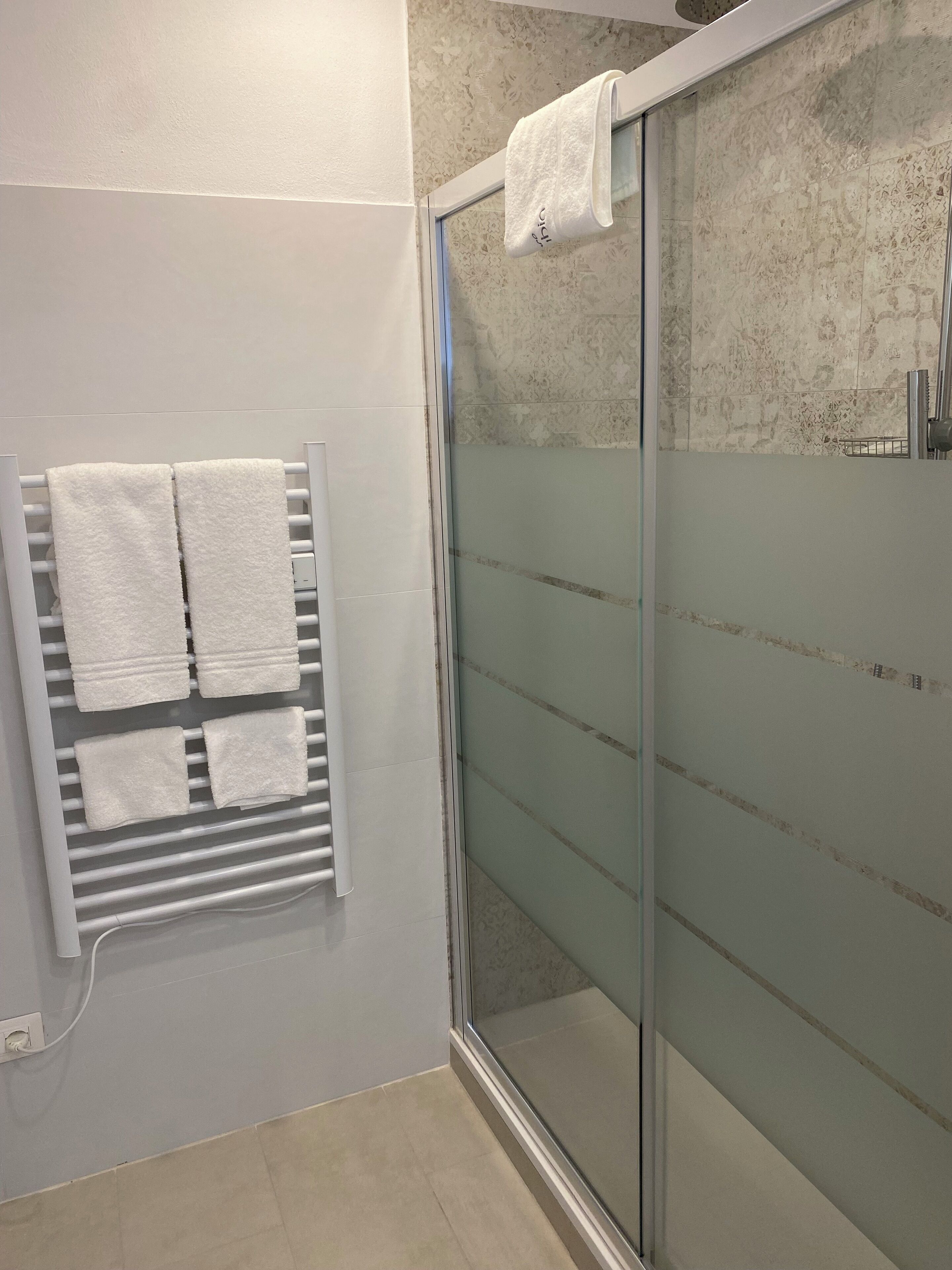 A bathroom with a glass shower enclosure, a towel rack with multiple white towels, and a tiled floor.
