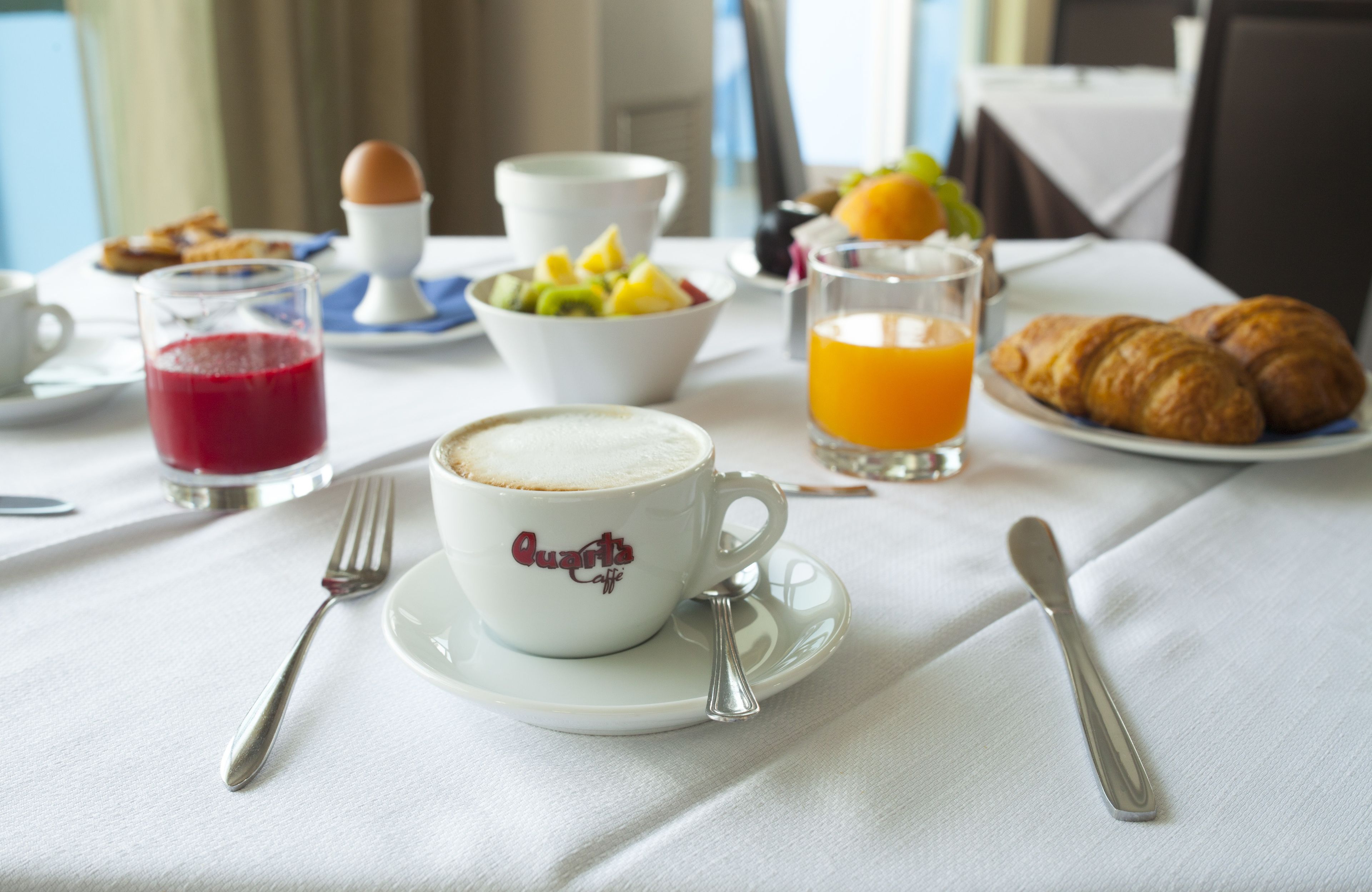 Una mesa de desayuno con una taza de café, un vaso de zumo de naranja, croissants, un bol de fruta y una fuente de tostadas.