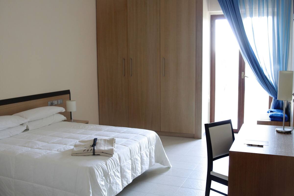 Una habitació d'hotel amb un llit gran, un armari de fusta, una taula de treball amb una cadira i una finestra amb cortines blaves.