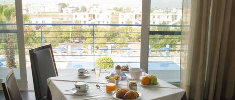 A breakfast table with croissants, fruit, and juice, set on a balcony with a view of buildings and a pool.