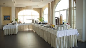 Una llarga taula de banquets preparada amb diferents plats i begudes, grans finestres amb cortines i un quadre a la paret.