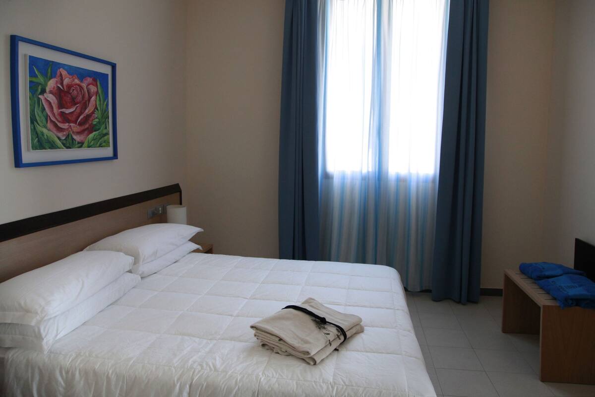 Habitació d'hotel amb llit, una taula de nit, una banca, un quadre i una finestra amb cortines.