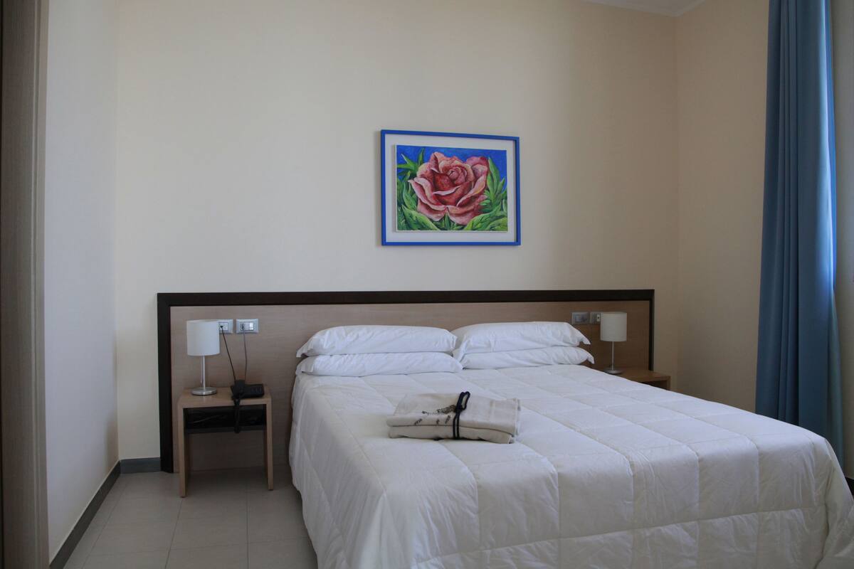Habitació d'hotel amb un llit, una nitòria amb una làmpada, un quadre d'una rosa a la paret i una cortina blava.