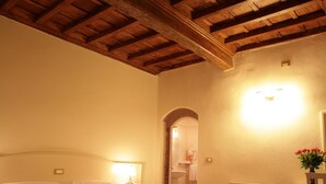 A bedroom with a wooden ceiling, a bed with a red quilt, a bedside table with a lamp, and a small desk with a chair. - Hotel Villa S Michele (Carmignano)
