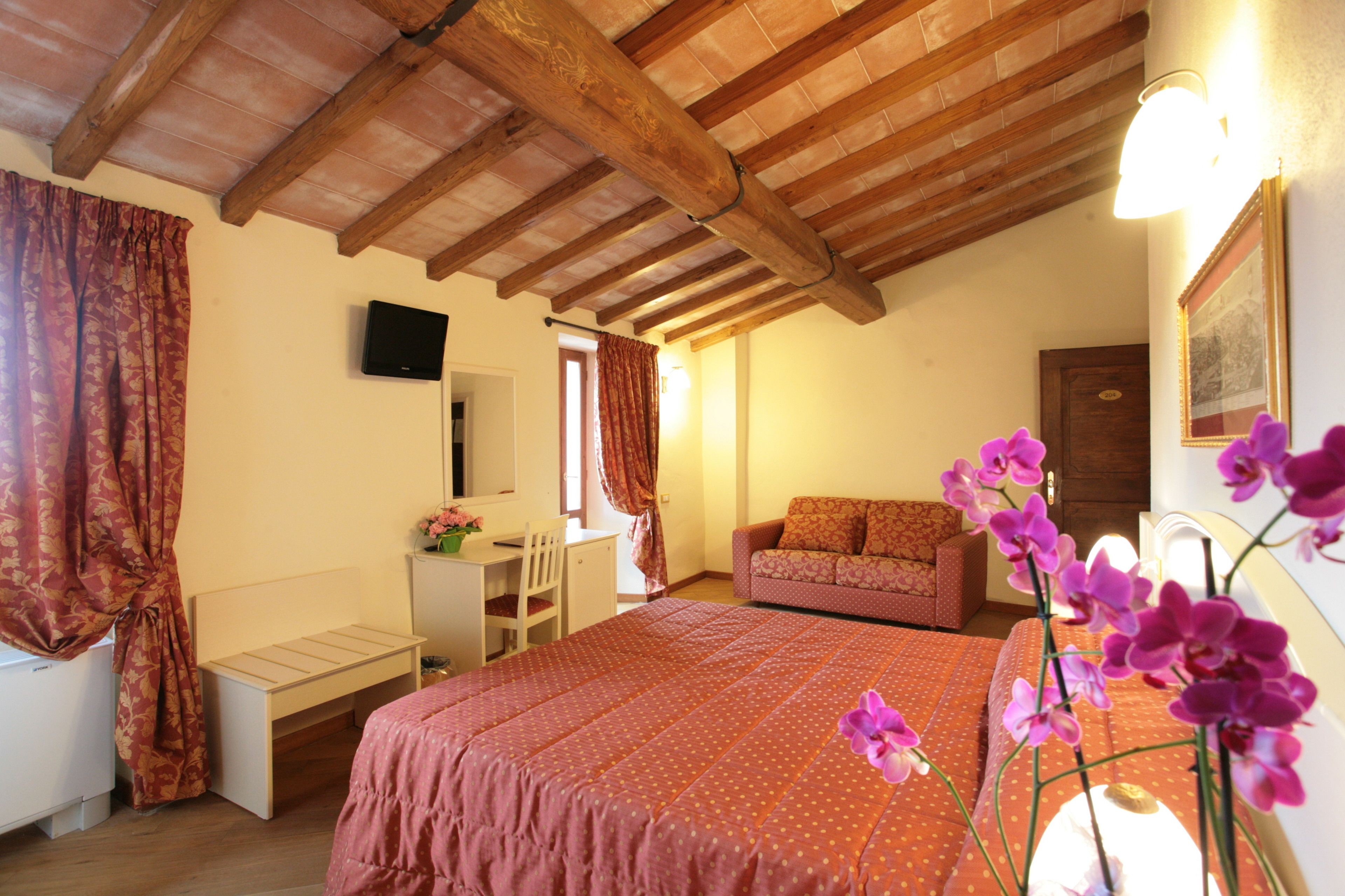 Una camera da letto con soffitto in legno, un letto con copripiumino rosa, una scrivania con sedia, un divano e una TV a parete.