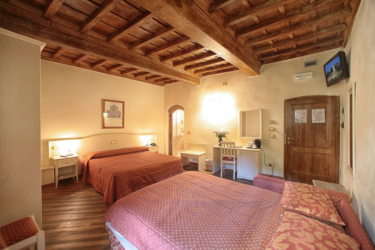 A hotel room with two beds, wooden ceiling beams, a desk with a chair, a mirror, and a door with papers on it.