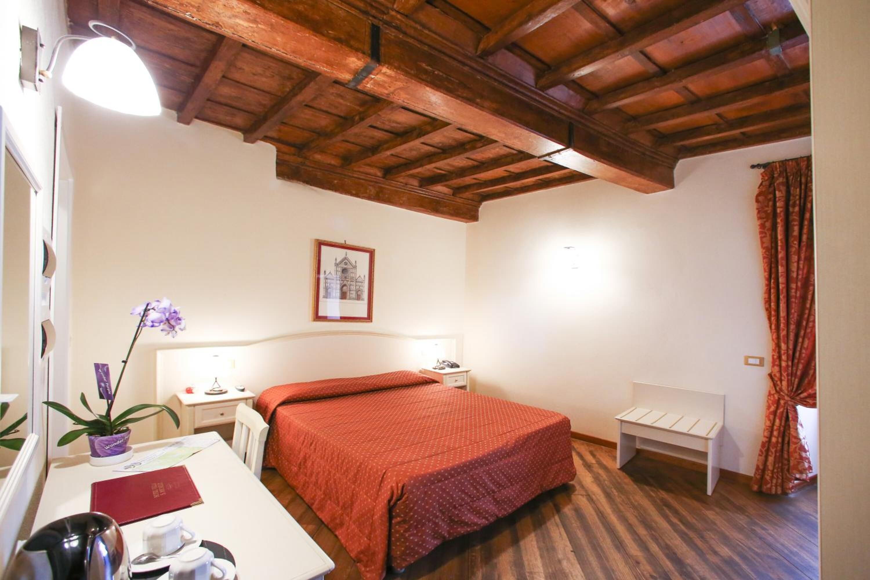 Ein Schlafzimmer mit Holzbalkendecke, einem Bett mit roter Decke, einem Nachttisch mit Lampe, einem kleinen Schreibtisch mit Pflanze und einem gerahmten Bild an der Wand.