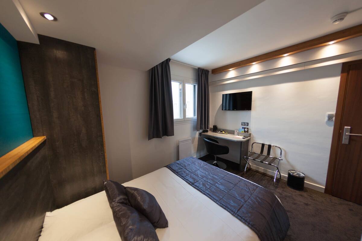 A modern hotel room with a bed, a desk, a chair, a television, and a window with curtains.