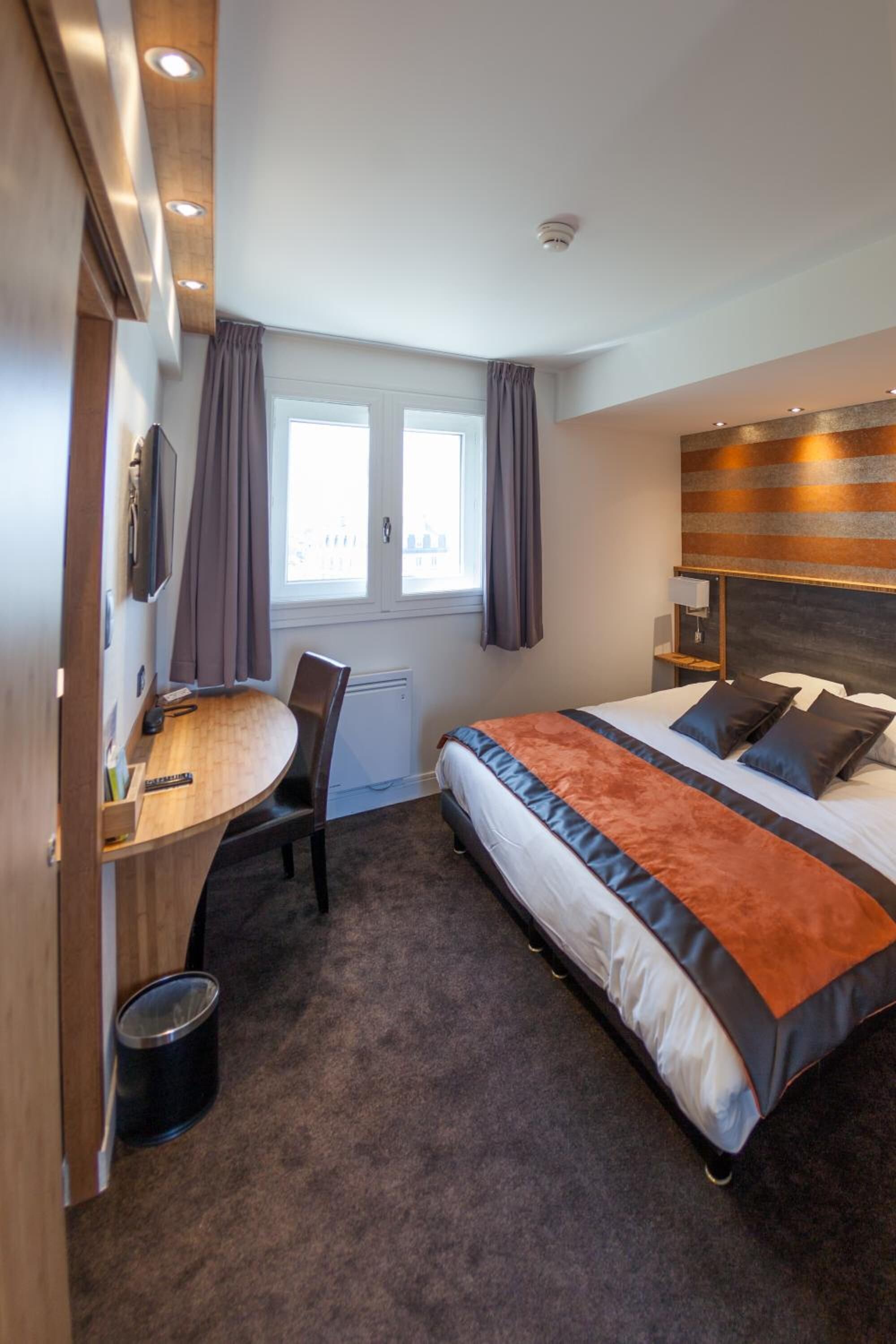 Ein Hotelzimmer mit Bett, Schreibtisch, Stuhl, einem Fenster mit Vorhängen und einem an der Wand befestigten Fernseher.